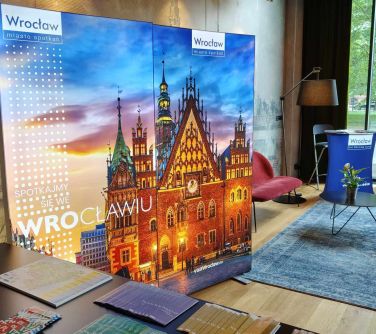 Pomieszczenie. Stolik, dywan, lampa, baner. Po lewej na ścianie oparty duży widok zamku we Wrocławiu