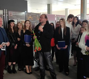 Pomieszczenie. Grupa osób, przed nimi mężczyzna z małym bukietem czerwonych kwiatów, przemawia do mikrofonu. W tle obrazy