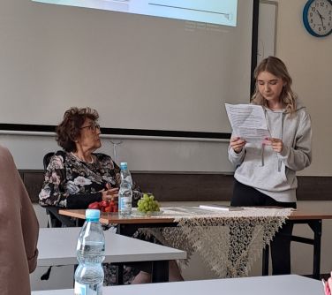 Sala lekcyjna. Starsza kobieta siedzi przy pierwszej ławce, patrzy na stojącą i czytającą z kartki dziewczynę. W tle na...