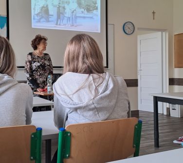 Sala lekcyjna. Przy ławkach siedzi czworo nastolatków. Na przodzie przy ławce siedzi kobieta, duga stoi przodem i opowiada