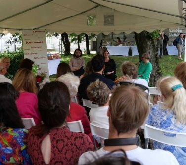 Na dworze, w ciągu dnia. Pod białym namiotem na krzesłach siedza słuchacze. Na przodzie trzech prelegentów