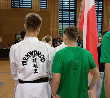 Aula sportowa. Dwóch nastolatków stoi tyłem. Jeden w białym strojy z napisem taekwon-do, drugi w zielonek koszulce z...