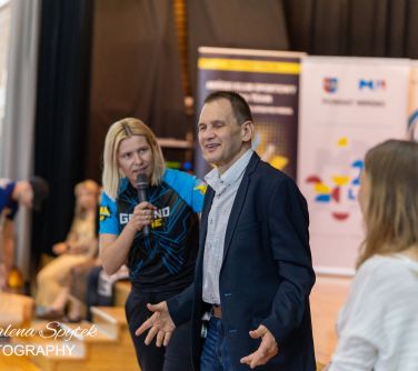 Hala sportowa. Kobieta przemawia do mikrofonu, patrząć na uśmiechniętego mężczyznę