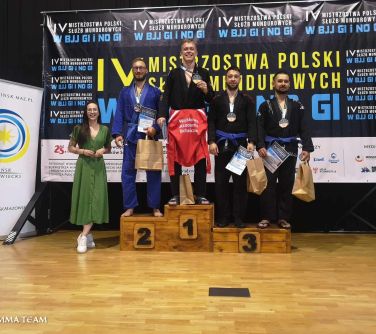 Hala sportowa. Na podium cztery osoby. Po lewej uśmiechnięta kobieta w długiej zielonej sukience i białych trampkach
