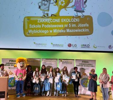Aula. Ma pzodzie przed banerami dziewięć osób po lewej osoba pzebrana za rysunkowego chłopca. Na górze na dużym ekranie...