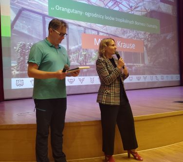 Aula. Przed sceną mężczyzna i kobieta. Kobieta przemawia