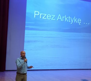Oświetlona aula. Po lewej mężczyzna przemawia stojąc poniżej sceny.