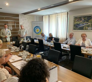 Sala konferencyjna. Stoły ustawione w litere U. Naokoło siedzą osoby. Dwóch mężczyzn stoi, jeden przemawia.