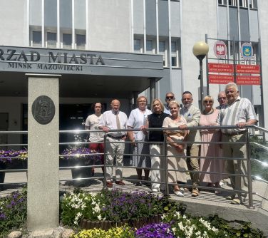 Na dworze, przed budynkiem urzędu. Dziesięc osób pozuje stojąc za metalową barierką