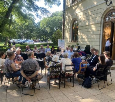 Na dworze. Na małym placu, na krzesłach wśród drzew i pałacu siedzą osoby. Rozmawiają