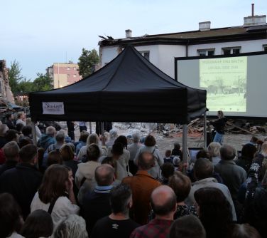 Za dnia duża grupa osób ogląda film wyświetlany na dużym ekranie. Po środku czarny namiot