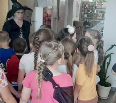 Wnętrze biblioteki. Kobieta w stroju czarownicy mówi do dzieci stojących naprzeciwko niej.