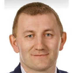 Zdjęcie portretowe. Mężczyzna uśmiecha się. Ma na sobie białą koszulę, granatową marynarkę i krawat.