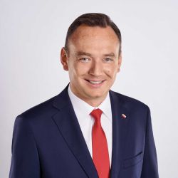Zdjęcie portretowe. Mężczyzna się uśmiecha. Ma na sobie białą koszulę, czerwony krawat i granatową marynarkę.