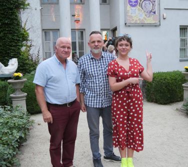 Ogród pałacowy za dnia. Przed pałacem trzy osoby. Kobieta palcami wskazuje za siebie na pracę powieszoną na ścianie.