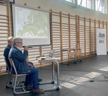 Na hali sportowej za dnia. Z lewej przy drabinkach na dwóch krzesłach siedzą mężczyźni. Jeden przemawia. przed nimi mały...