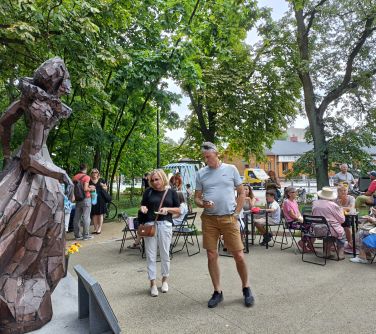 Skwer, w ciągu dnia. Przy stolikach na krzeslach siedzą osoby. Kobieta i mężczyzna ogląają posąg mosiężny kobiety