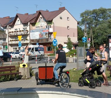 Na placu na zewnątrz osoby. Po środku mężczyzna jedzie rowerem z bagażnikiem z przodu na książki. W tle budynki