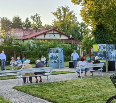 Ogród. Białe ławeczki. Między alejkami wystawa. Na przodzie mężczyzna przemawia do mikrofonu