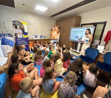 Jasne pomieszczenie. Dzieci siedzą na piankowych matach. na przodzie kobieta stoi przy dużej tablicy interaktywnej