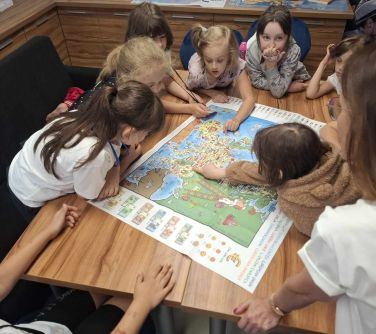 Jasne pomieszczenie. Przy stole na krzesłach siedzą dzieci oglądają mapę