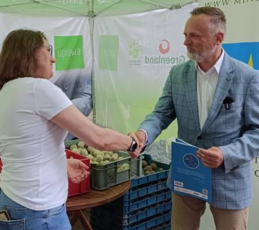 Na dworze, pod jasnym namiotem. Uśmiechnięty mężczyzna ściska dłoń kobiety. W drugiej dłoni trzyma teczkę