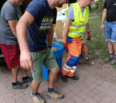 Na dworze, alejka w parku. Dwóch mężczyzn niesie plastikową skrzynkę wypełnioną kulkami z gliny. Naokoło osoby