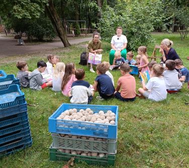 Park. Na trawie siedzi grupa dzieci. Przed nimi dwie kobiety opowiadają. Na przodzie ustawione na sobie skrzynki z...