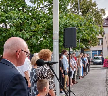 Na zewnątrz. Ludzie w rzędzie stoją. Na pierwszym planie mężczyzna mówi do mikrofonu.