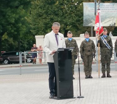 Na zewnątrz. Mężczyzna w białej marynarce wygłasza przemówiwenie. Stoi przy mównicy. w tle stoją żołnierze w mundurach.