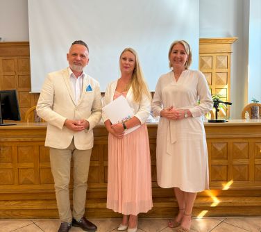 Oświetlona aula. Na tle drewnanego wysokiego stoły pozują trzy osoby. Po środku kobieta trzyma dyplom w dłoniach