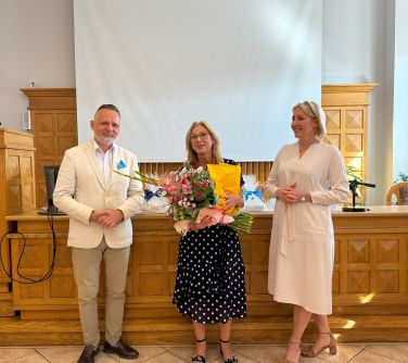 Oświetlona aula. Na tle drewnanego wysokiego stoły pozują trzy osoby. Po środku kobieta trzyma bukiet kwiatów