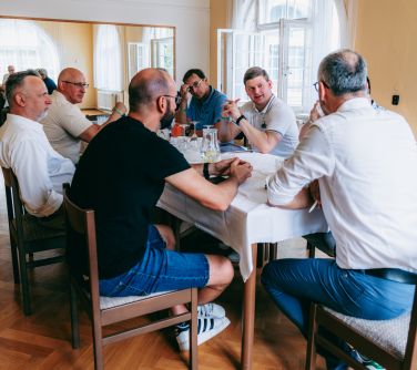 Pomieszczenie. Przy stole na krzesłach siedzi sześciu mężczyzn. Na ścianie duże lustro