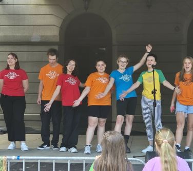 Dzień. Na scenie grupa osób ubranych w kolorowe podkoszulki