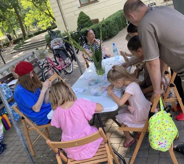 Dzień. Przy stole naokoło na krzesłach siedzą osoby.