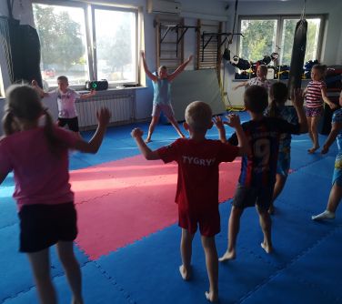 Na sali ćwiczeniowej grupa dzieci ćwiczy na piankowych matach. W tle okna i drążki