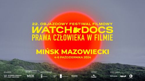 Na tle nieba i na dole zieleni po środku duże czerwone koło. Na nim zółte napisy