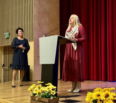 Sala koncertowa. Na scenie dwie kobiety, jedna przemawia przed mównicą. Na przodzie żółte kwiaty