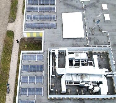 Dwór. Dach budynku z instalacją fotowoltaiczną. Po lewej dwa pasy trawy i chodnik po którym idą dwie osoby, jedna z wózkiem.