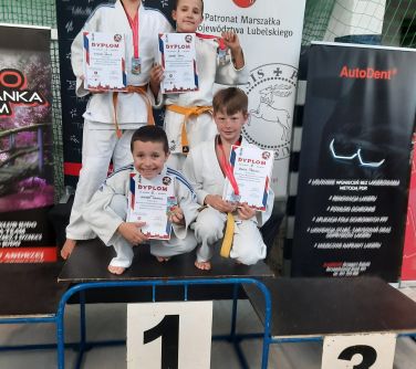 Pomieszczeie. Czwórka chłopców w białych kimonach pozuje na 1 miejscu podium. Dwóch z przodu kuca. W rękach trzymają dyplomy