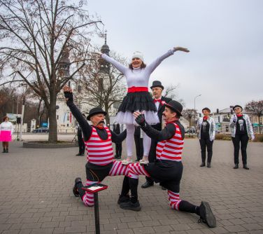Na dworze, dzień. Grupa seniorów w układzie tanecznym. Dwóch mężczyzn klęczy, seniorka na ich kolanach