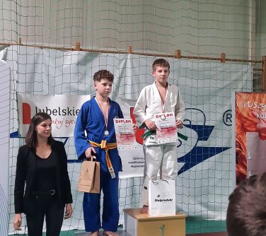 Hala sportowa. Na podium dwóch chłopców z dyplomami. Po lewej ubrana na czarno kobieta