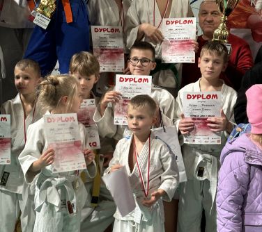 Grupa dzieci w białych kimonach pozuje do z dyplomami. Miedzy nimi mężczyzna z pucharem
