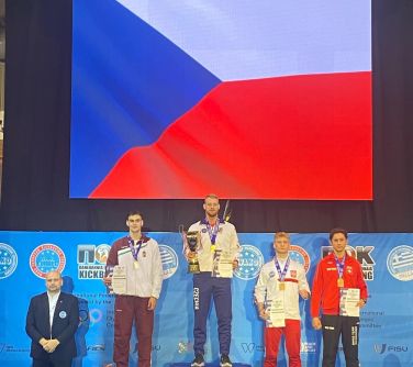 Na podium cztery osoby. Po lewej mężczyzna w ciemnym garniturze. W tle biało-niebiesko-czerwona flaga czech