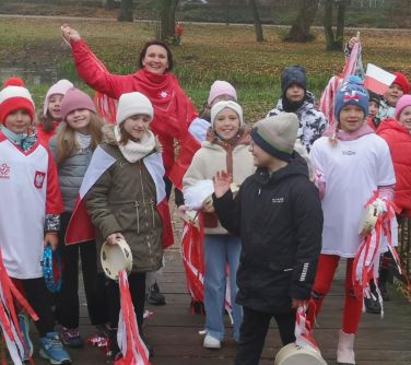 Na dworze w pochmurny dzień. Grupa uśmiechniętych dzieci oraz kobieta ubrana w czerwoną kurtkę stoją na kładce w parku...
