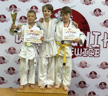Hala sportowa. Trzech chłopców stoi na podium trzymając się za ramiona. Dwóch trzyma dyplom, a jeden puchar