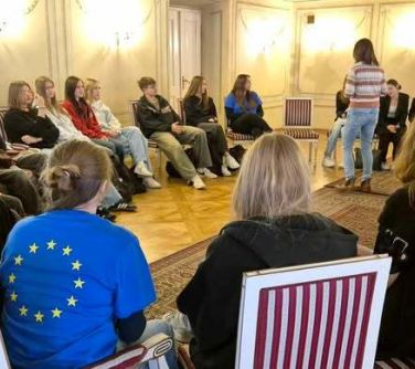 Oświetlona sala w klepce z dywanem. Naokoło na krzeslach siedzi  młodzież. W środku jedna kobieta stoi tyłem