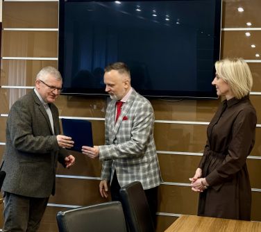 Pomieszczenie. Przy stole trzy osoby. Mężczyzna wręcza mężczyźnie pismo