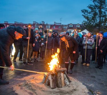 Na dworze. lekki zmierzch. Naokoło rrozpalonego ogniska zgromadzone osoby. Po lewej mężczyzna ciemnej kurtce podpala od...