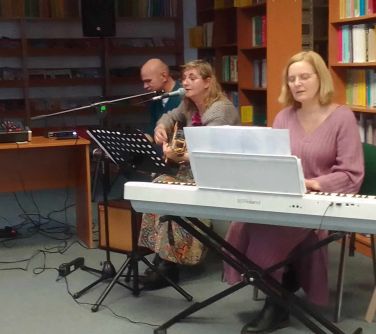Pomieszczenie. Trzy osoby siedzą na krzeslach i grają na instrumentach. W tle półki z książkami